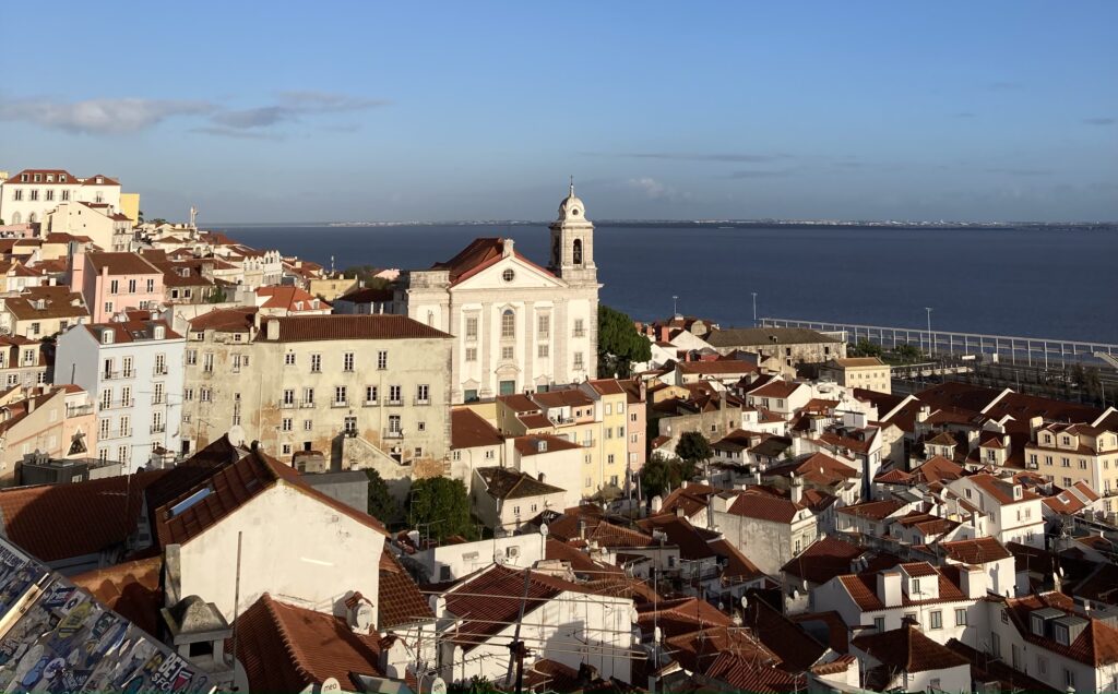 Image of Lisbon, Portugal taken from the Santa Lucia miradouro. We can see the different buildings, a church and the ocean in the background.