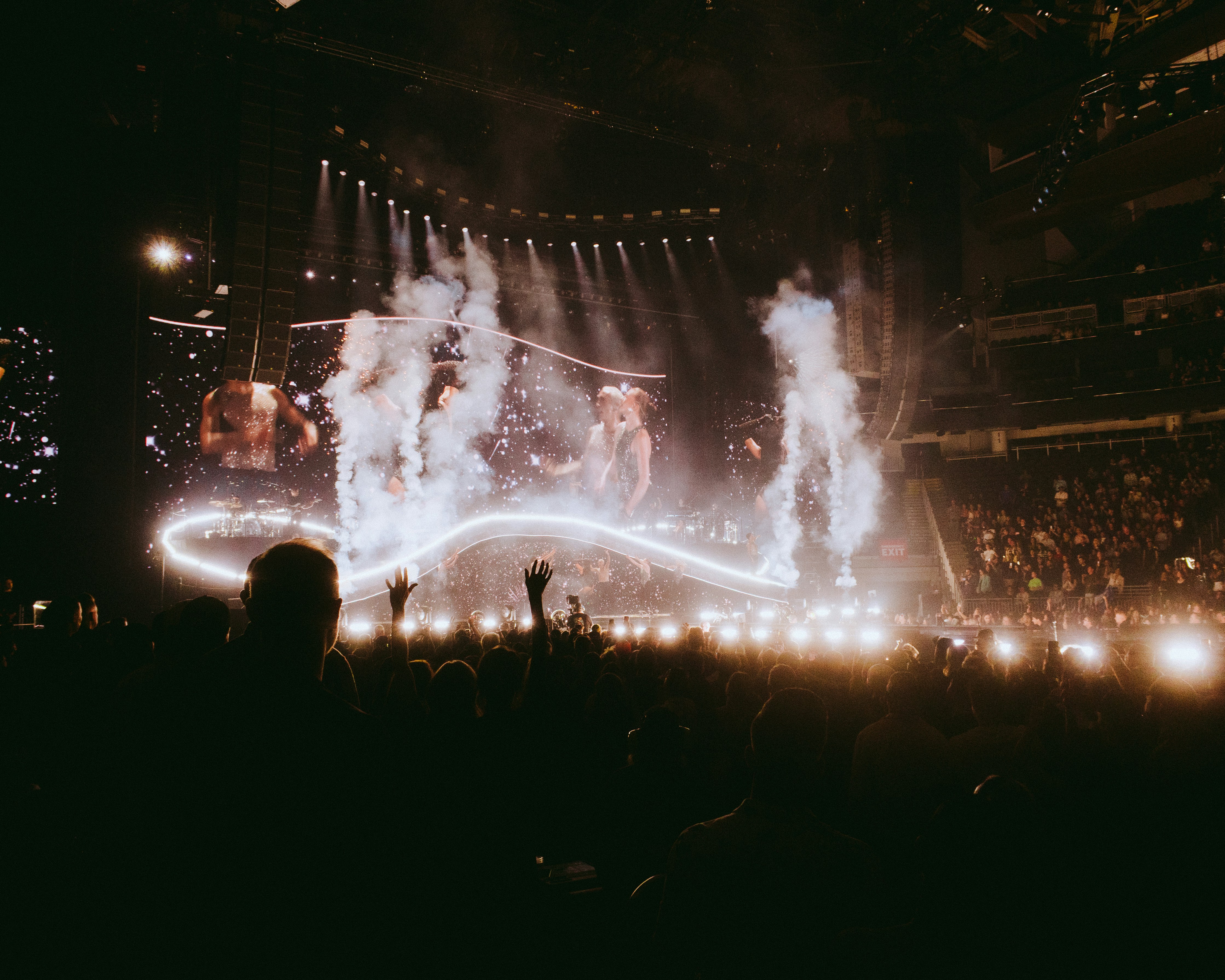 Epic concert crowd with hands up, dramatic stage lights and smoke effects capturing live music immersion from spectator view